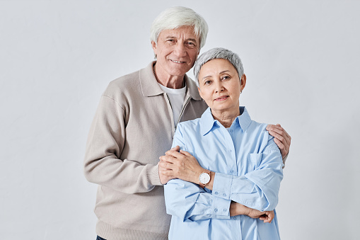 Waist up shot of smiling white-haired Caucasian senior man hugging shoulders of Asian senior woman while both looking at camera