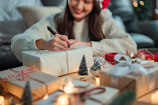 Hands of woman writing greeting card on Christmas gift, Female preparing present gift box on the table, Christmas eve preparation