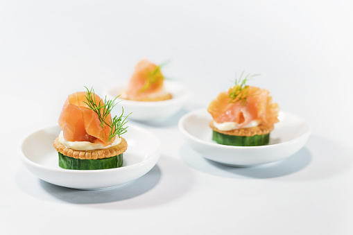 Canapes as party finger food for the cold buffet made from cucumber, crackers, cream cheese, smoked salmon and dill on small white plates, copy space, selected focus, narrow depth of field