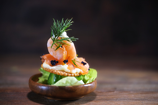 Shrimp, salmon and caviar on a cracker with mayonnaise in a small wooden bowl on a dark rustic background, snack for a holiday party like Christmas or New Year, copy space, selected focus
