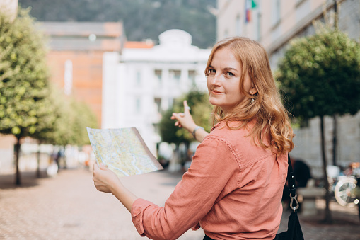 Portrait beautiful blonde woman with paper map in street. Happy tourist travels in Europe. Vacation concept by exploring interesting places to travel. Women Searching locations at summer day.