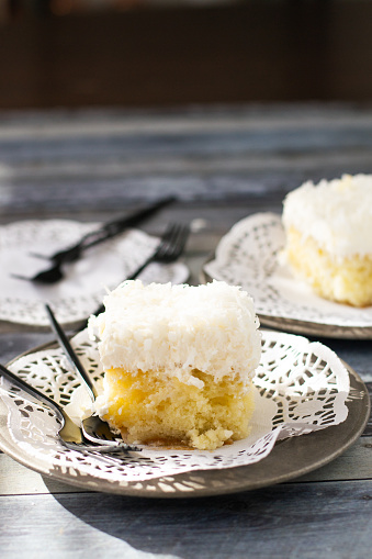 Two pieces of Coconut Cream Poke Cake, vanilla cake with whipped cream and shredded coconut, on a white doille with two black forks laid across a grey plate on a blue grey background with black forks in the background.