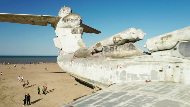 a white ekranoplane is standing on the seashore, on a sandy beach, tourists are walking around, a drone shot, slow motion, flying around the plane, airplane details