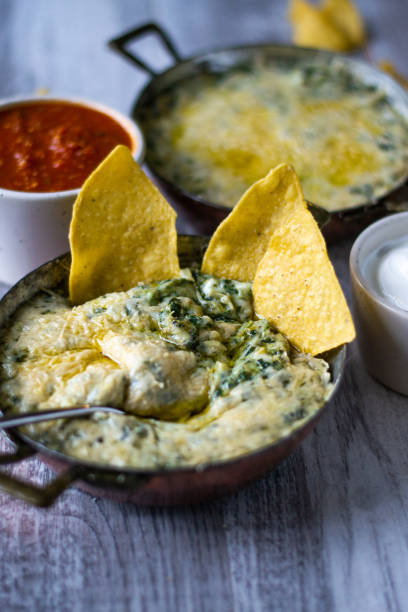 Spinach and Artichoke Dip with two tortilla chips sticking into it inside a copper and metal bowl with two handles on a light grey background with another metal bowl of dip in the background and two small cream bowls of salsa and sour cream.