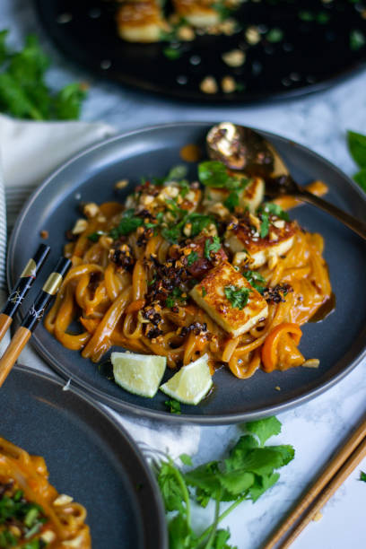 istock Plated sauced PB noodles with crispy halloumi; garnished with cilantro, peanuts, red pepper flakes served with chopsticks and a gold spoon on the side of the plate. 1877891271
