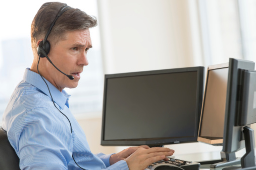 Young man with headphones, at computer, working in tech support or customer care. Happy, smiling male professional call center agent in office. IT technician, telemarketer or online gamer on laptop.