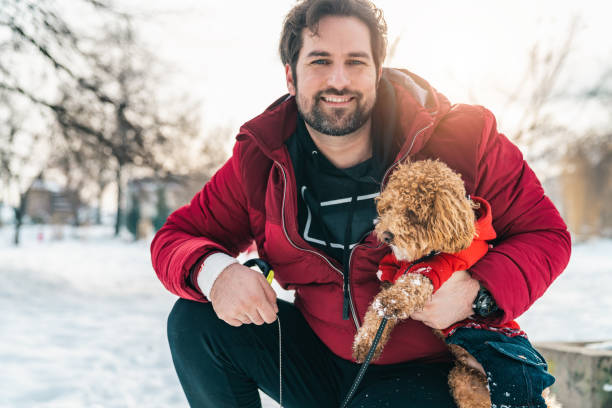Handsome male pet owner looking at camera while holding his poodle dog and posing for a photo in the snow. Happy smiling man with toy poodle outdoors on sunny winter day.