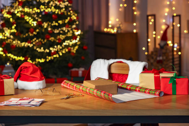 istock Santa's Claus workplace. Gift boxes, wrapping paper, letters on table and costume in room with Christmas tree 1878035708