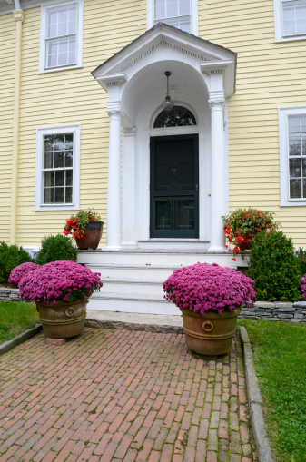 yellow multi-story house with a portico, brick sidewalk and flowers with pink flowers
