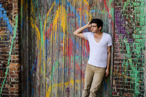 A handsome young man in a v-necked t-shirt leaning against a old, wooden, paint-splattered door and brick wall. Adobe RGB color space.