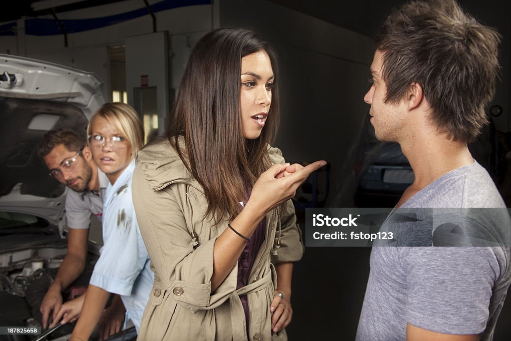 Service Industry: Couple argues over vehicle repairs. Couple arguing over vehicle repairs. Mechanics watch in background. 20-29 Years Stock Photo