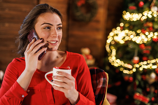 Portrait of joyful young White woman sitting at her desk, enjoying a cup of coffee and talking on the phone during work break. Christmas tree glistening in the background.
