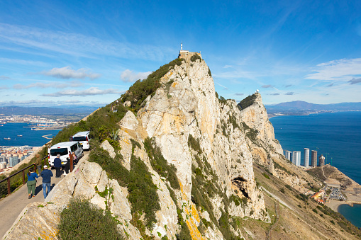 Gibraltar - 11th November 2023: Tourist mini-buses on St Michael Road looking towards Signal Hill. Some tourists are walking on the road, and there is a Barbary Macaque on top of the rear vehicle. On the right is part of the city of Gibraltar and the bay of Gibraltar looking back to Algeciras in Spain. On the left is Catalan Bay, Hassan Centenary Terraces and a Spanish sierra.