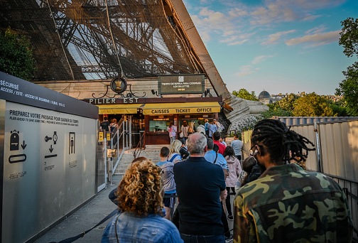 People queuing to buy tickets and access the Eiffel Tower. The warm light of late afternoon illuminates it fully.