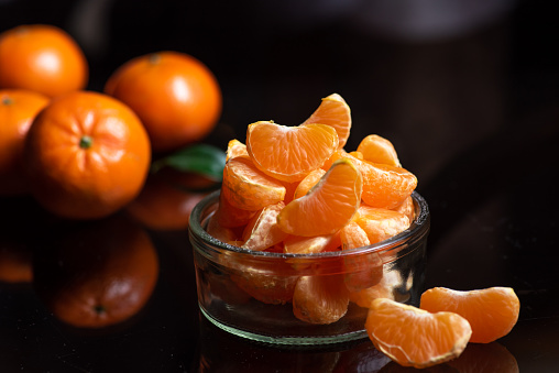 Slices of mandarins in a glass bowl and whole mandarins on a glass table, with reflection