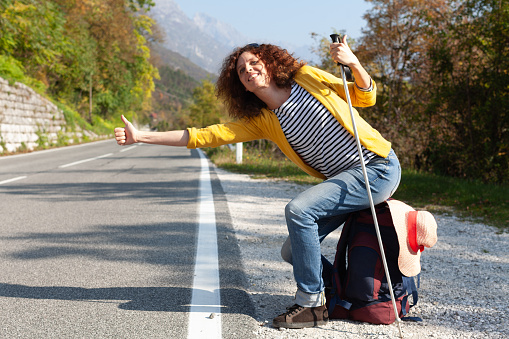 Cheerful Curly Hair Mid Adult Caucasian Woman Backpacker Hitchhiking on a Road