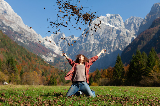 Cheerful Young Woman Kneeling on Meadow and Enjoy Throwing Autumn Leafs in Air on a Beautiful Mountains  Background of Her First Day of Vacations
