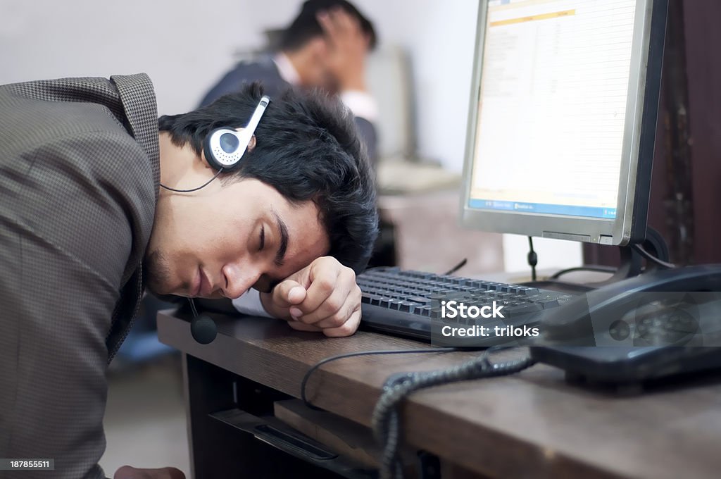 Business executive sleeping at his desk with headphones on Disappointed young customer service operator wearing headset with desktop pc and sleeping in an office. Adult Stock Photo