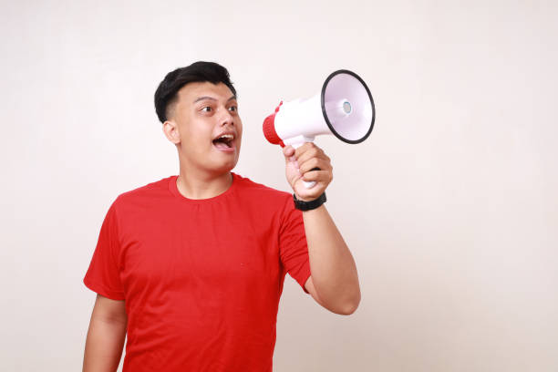 istock Funny young asian man announcing shouting to sideways using megaphone. Isolated on white 1878581432