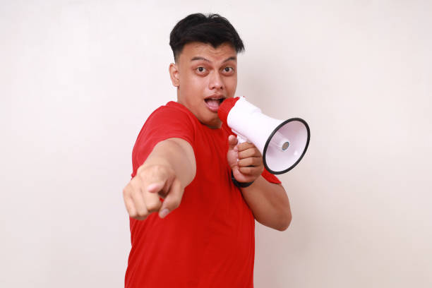 Handsome Asian young man pointing at you while speaking holding megaphone. Isolated on white