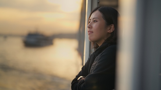 An Asian female tourist is traveling alone on a ferry during sunset.