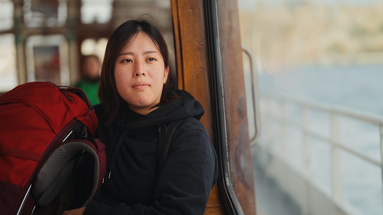 An Asian female tourist is sitting inside of a ferry and traveling alone.