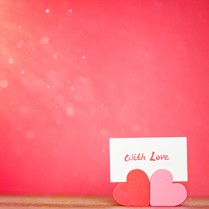 Hearts holding a sign “with love”. Red background with copy space. For care concepts, romantic events such as Valentine’s Day and anniversaries.