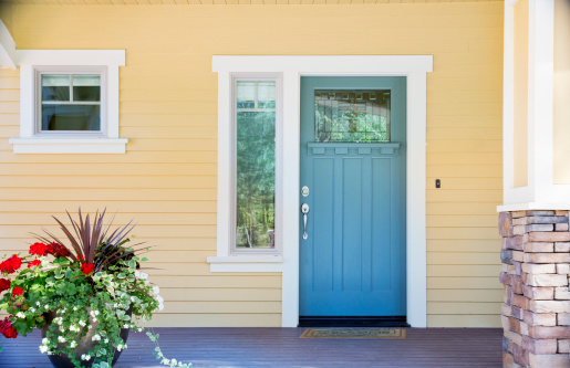 A front entrance of a home with a blue door, yellow siding, and a flowerpot in daytime.