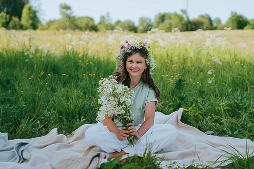 Cheerful brunette schoolgirl in casual sitting on plaid at meadow, with wreath on head holds bouquet of wildflowers toothy smiles looks at camera. Caucasian teen girl enjoying nature on vacations.