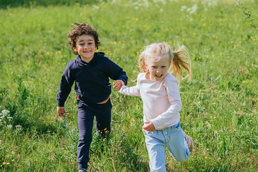 Cheerful  brunette curly hispanic boy running at meadow with blonde caucasian little girl smiles wide on sunny summer day. Childhood outdoor leisure. Pretty Swedish girl holds  brothers hand.