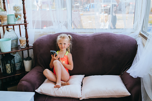 Curious young girl sitting on a sofa, holding a smartphone, by a window adorned with tulle curtains and home decor