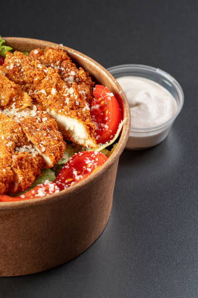 Snittel Caesar Salad in a paper takeaway container with sauce on a black background. Healthy eating concept, restaurant food delivery. View from above. Free space for your text.