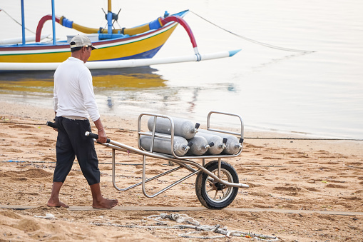 Bali, Indonesia - October 23, 2023:  Male worker is carrying oxygen cylinders for divers on a wheelbarrow at Sanur Beach, Bali, Indonesia during the day. Concept for scuba diving and labor day.