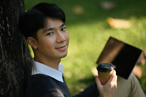 Close-up image with Young Asian university student man sitting under the tree while holding a coffee and tablet.
