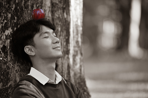 A Young Asian man with a Red apple on his head sitting under the tree, sepia-toned.