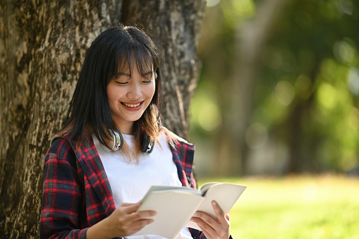 A Happy Young Asian Girl enjoying a book under the tree in the outdoor park.