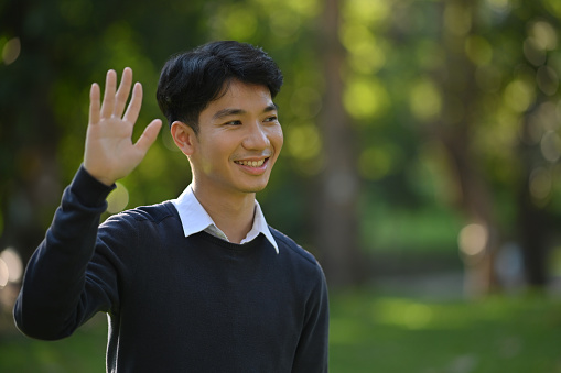 A Young Asian university student man waving a hand to greet his friend in the park.