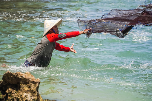 Yogyakarta, Indonesia - August 23, 2023: A male fisherman wearing woven bamboo conical hat is catching anchovies using a net on a rocky beach during sunny day at Ngrenehan Beach, Gunungkidul, Jogja. Throwing Fishing Net by Hands.