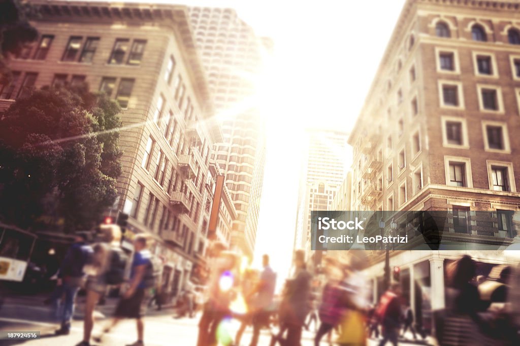 People on street of downtown San Fransisco People walking across a street.  The street runs between some tall buildings with even taller buildings in the background.  The sun is shining at the end of the street, casting bright light on the people.  There is a tree next to the building on the left. People Stock Photo