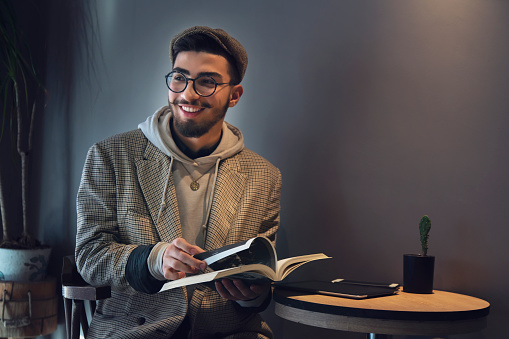 Young handsome man in eyewear spending free time and reading interesting book in coffee shop, serious middle eastern man student spending free time on education in coffee shop.