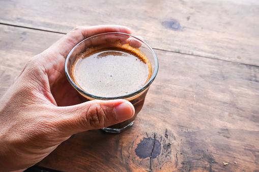 Someone is holding a cup of hot coffee on a wooden table. Black coffee with crema foam on the surface of the drink. Empty blank text copy space.