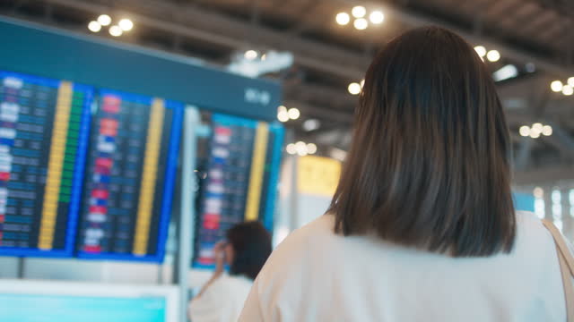 Young Asian female passenger with luggage holding smartphone, looking arrival departure board, checking flight information board, walking and standing in international airport, Happy woman waiting for travel trip