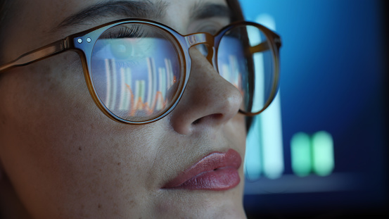 Close up of a young woman data graph on a computer screen reflecting in her spectacles.