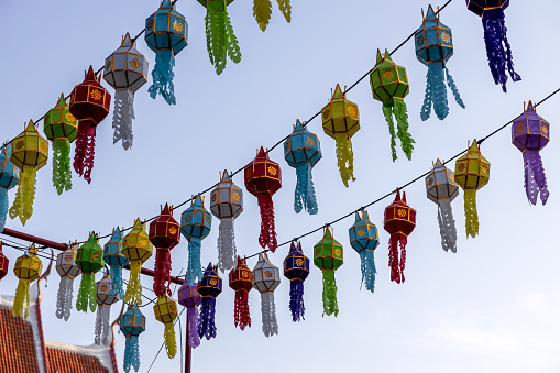 A view from below of colorful lantern flags hanging on strings of lights with the blue sky in the background during the day before the Thai Loy Krathong festival.
