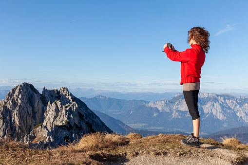 Curly Hair Young Woman Taking a Photograph with Her Smart Phone from a High Viewpoint in High Mountains