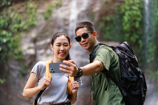 Happy young couple taking selfie with smartphone in front of tropical waterfalls in summer vacation.