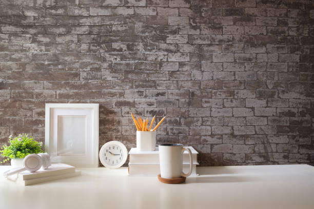 istock Home office interior. Picture frame, coffee cup, notebook and pencil holder on white table against brick wall. Copy space. 1879748163