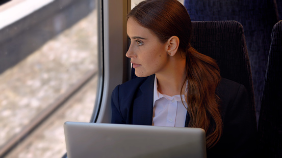 A young woman working at her laptop on a train journey, she takes a moment to think and look out of the window.