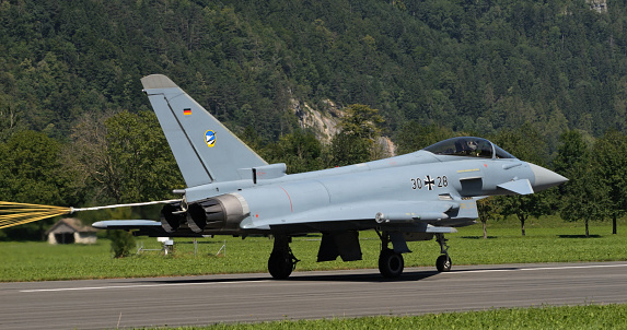 Mollis Switzerland August 19 2023: Fighter Jet Landing with Brake Parachute in Mountain Air Base Runway in a Sunny Day. Copy Space. Eurofighter Typhoon of German Air Force.