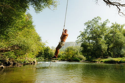 Photo of a man enjoying a scorching summer day by the river, joyfully swinging on a rope resembling a liana.
